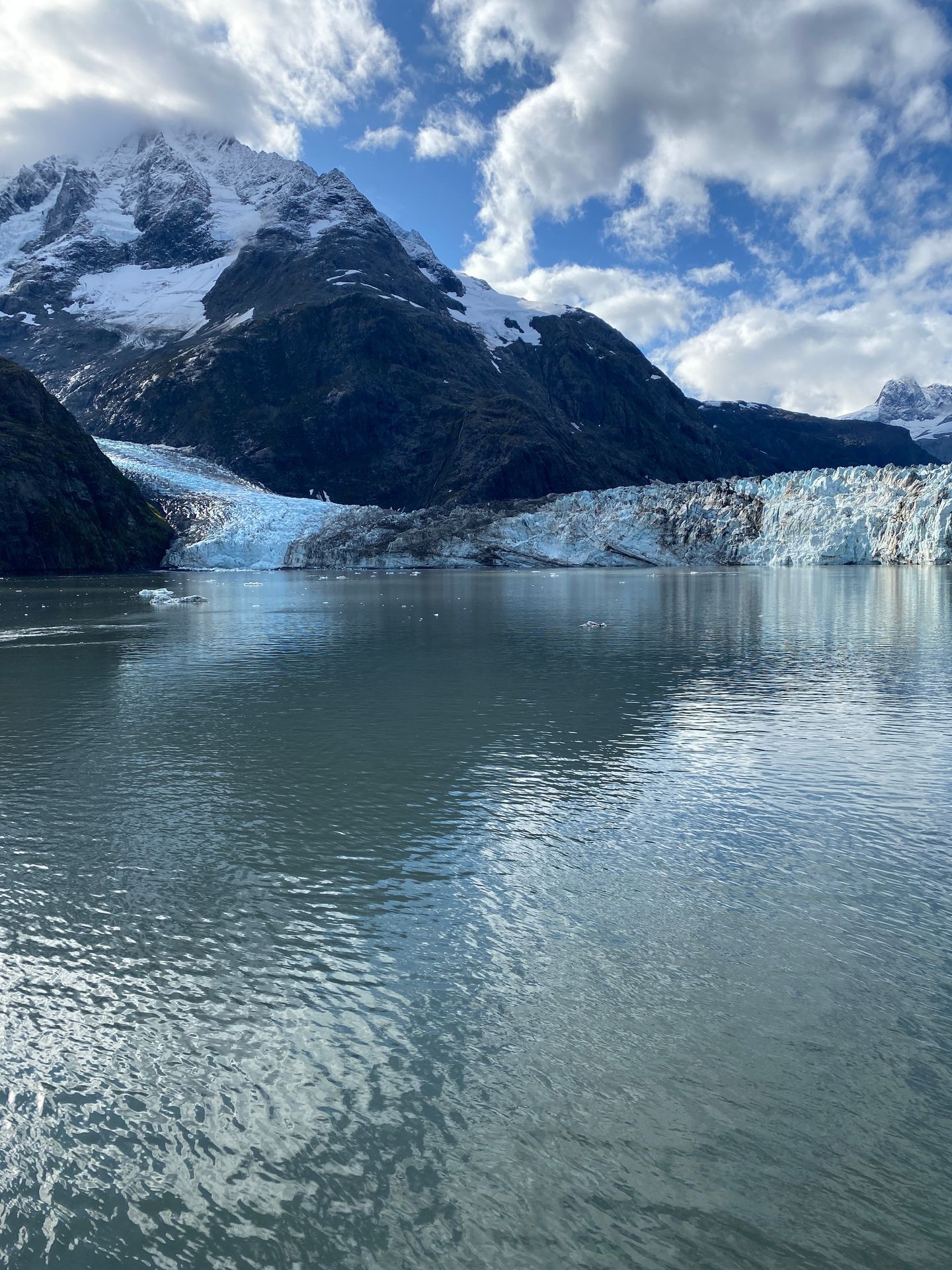 A breathtaking Alaskan glacier meets the icy waters below, framed by towering, snow-capped mountains under a bright blue sky with scattered clouds.